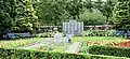 Photo d'un jardin avec des buissons bien taillés et des allées de fleurs de chaque côté. Au milieu se trouve des pierres commémoratives et dans le fond un monument en trois parties avec l'inscription du nom des victimes.