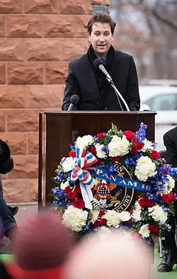 Photo d'un homme se tenant derrière un pupitre avec un micro et prononçant un discours. Il se tient juste devant un monument commémoratif en pierre rouge et une couronne de fleurs est fixée devant le pupitre.