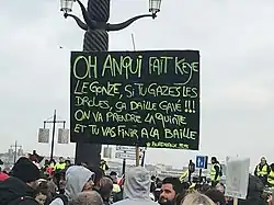 Photographie de manifestants portant un panneau noir avec une inscription jaune en bordeluche.