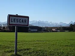 Photographie en couleurs d'un panneau avec l'inscription " Lescar", en fond des montagnes enneigées.