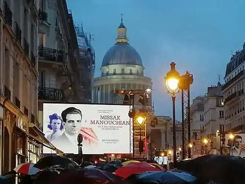 Vue du Panthéon en soirée. Au premier plan, une foule de parapluies surmontée par un panneau d'affichage montre Missak et Mélinée.