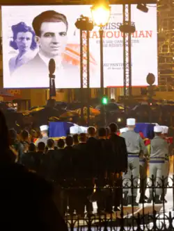 Photographie d'un cortège funéraire. Des militaires portent deux cercueils, entourés d'une foule protégée par des parapluies. En arrière-plan, un panneau d'affichage montre Missak et Mélinée.