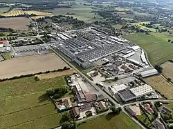 Vue aérienne montrant une vue d'ensemble d'une zone industrielle entourée de paysages ruraux. Au centre de l'image, on observe le complexe industriel de Lamberet comprenant plusieurs bâtiments d'usines et des entrepôts.