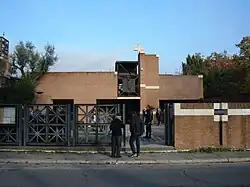 Une église à l'architecture moderne, en brique. Elle est surmontée d'une croix carrée et accompagnée d'un portail en fer forgé qui donne sur la rue.