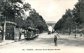 Photo noir et blanc d'une grande artère bordée d'arbres avec piétons, tramway, voitures à cheval, édicule à marquise ronde et au fond, l'arc de triomphe