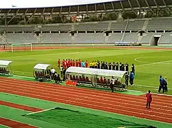 Photographie d'un stade de football, avec plusieurs joueurs sur la pelouse, alignés et de dos.