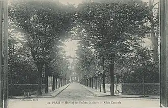 Photographie en noir et blanc d'une allée bordée d'arbres au bout de laquelle on aperçoit des bâtiments