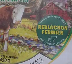 Gros plan sur la pastille verte et jaune figurant sur l'emballage d'un reblochon fermier, à l'arrière plan figue sur l'emballage le dessin d'un chalet et d'une vache de race montbéliarde.