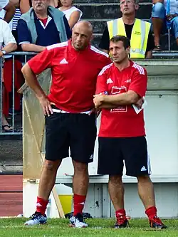 Deux hommes observent un match de rugby depuis le bord du terrain.