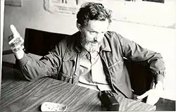 Un homme barbu, attablé dans un bar, lève la main droite.