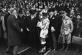Homme à barbe blanche portant un haut-de-forme remettant une coupe à un footballeur, avec une foule autour et derrière lui