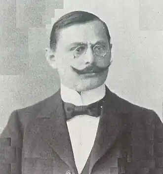 Portrait en noir et blanc d'un homme aux cheveux bruns et courts, habillé d'un costume sombre, arborant un pince-nez, une moustache en guidon et un nœud-papillon.