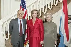 Ces trois personnes posent devant le drapeau américain et le drapeau du Luxembourg, le tout devant un escalier blanc.