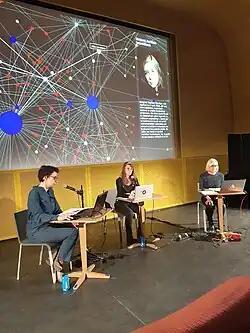 Projection d'une galaxie de liens en arrière plan, 3&nbsp;femmes réparties sur une scène lisant du texte à tours de rôle sur leur ordinateur portable, dans un micro
