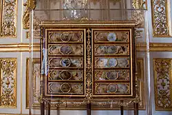 Cabinet-médailler_petit appartement du Roi Versailles; acquis à titre personnel par Louis XVI peu de temps avant la Révolution. Exécuté en bois d'acajou et en bronze doré, il présente un surprenant décor composé de panneaux de cire sur laquelle sont appliquées des végétaux, des plumes d'oiseaux et des ailes de papillon, ce qui en fait une véritable rareté, digne d'un cabinet de curiosité. Aliéné en 1796, le meuble fit retour à Versailles en 1986 et rejoignit le salon de musique, dans les cabinets du roi, où Louis XVI l'avait fait placé. Travaille ensuite sous l’Empire.