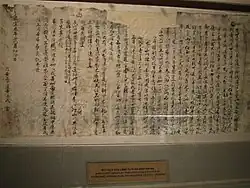 Morceau de papier abîmé sur lequel on lit des écritures en caractères chinois écrits à l'encre sombre. Le papier est encadré et exposé dans un musée.