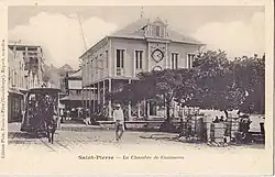 Photographie sépia d'un bâtiment à colonnades en bois donnant sur une vaste place arborée.