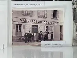 Photographie en noir et blanc. Devant un bâtiment à façade en pierre avec plusieurs fenêtres aux volets ouverts, au fronton duquel on lit « Manufacture de chemises », une quinzaine de personnes, dont une dizaine de femmes et 5-6 hommes, posent debout face à l'objectif. Tous et toutes ont des tenues sobres. Les femmes sont en robe longue ou pull et jupe longue et portent les cheveux courts ou en chignon. Les hommes sont en salopette et veste, ou en complet. Un homme sur la gauche porte un costume élégant avecu un chapeau sombre. Vers la droite, un autre porte un béret noir.