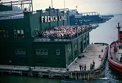 Un bâtiment au bout d'un quai, la façade marquée de l'inscription en grandes lettres « French Line ». Une foule attend sur la terrasse et salue l'arrivée du paquebot d'où la photo est prise.