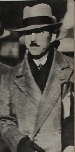 vue en plan rapproché d'un homme en manteau, foulard et chapeau, ganté, la cigarette aux doigts et la serviette sous le bras. Moustache et lèvres fines, l’œil perçant.