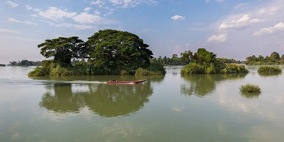 Pirogue filant sur le Mékong devant une petite île avec des arbres verts se reflétant dans l'eau, dans le Si Phan Don.