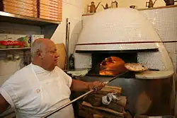 Photo d'un pizzaiolo en train de retirer une pizza d'un four à bois à l'aide d'un long manche. On voit le bois brûler à l'intérieur du four à travers son ouverture. L'extérieur du four est en céramique blanche, des buches de bois sont entassé au pied du four.