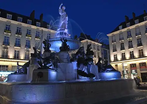 La fontaine, de nuit, éclairée en bleu sur fond d'immeubles éclairés en blanc.