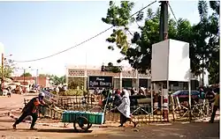 place en latérite avec quelques passants piétons, un arbre, un poteau électrique et une structure blanche sur le côté droit