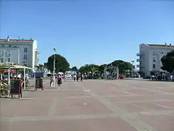 Photographie d'une vaste place vide au revêtement rosé quadrillé de pierres plates. Au fond la grille 1900 en fer forgé, les pins et palmiers du front de mer