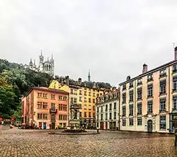 Vue d’une place avec des maisons colorées à trois étages sur deux côtés ; au fond, une colline boisée et une basilique.