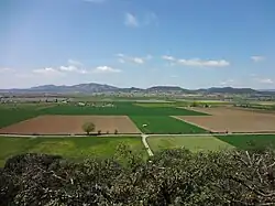 D'un point de vue en hauteur, on voit la plaine découpée en champs carrés de différentes couleurs. Au fond, petites montagnes dans le lointain, et ciel bleu moucheté de nuages.