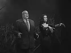 Un homme chauve en costume et une femme en robe et aux cheveux longs marchant de façon mécanique.