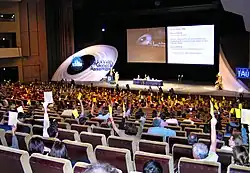 Photographie d'un amphithéâtre où de nombreuses personnes votent en levant la main avec un carton coloré.