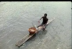 Photographie d'un homme, torse nu, assis à califourchon sur une planche en bois flottant sur l'eau. L'homme tient un filet contenant plusieurs poissons.