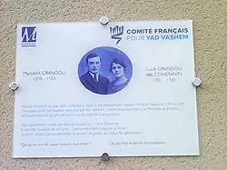 Plaque blanche portant en bleu la photo du couple Grandou jeunes, les logos de Mayrac et du Comité français Yad Vashem et une exhortation des passants à se souvenir.