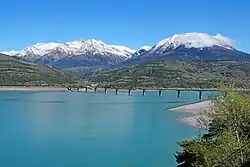 Photo du pont avec le lac au premier plan et des montagnes partiellement enneigées en arrière-plan.