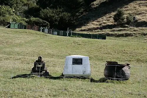 Monument de pierre entouré d'un canon ancien de navire et d'une sorte de chaudron métallique.