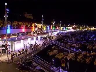 Photographie présentant un panorama du port la nuit: au premier plan des pontons de bois et des bateaux blancs, au second plan les quais illuminés où circulent des promeneurs.