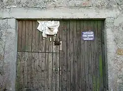 Une porte de grange en bois sombre située dans le hameau de Recoulette, fermée par des targettes en métal et entourée par des piliers et un linteau en béton. En haut à gauche, un morceau de tissu blanc plié dépasse de la porte. En haut à droite, un panonceau bleu avec un texte en blanc : "Département de la Lozère. Services vétérinaires. Étable indemne de tuberculose".