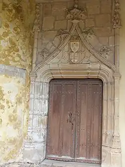 Photo couleur d'une porte en bois cloutée à deux ventaux. Les montants en pierre sont nervurés et le linteau est surmonté d'un arc brisé orné de blasons.