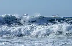 De grandes vagues se brisent sur le rivage, tandis que des navires de commerce passent au loin.