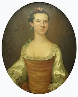 Portrait ovale d'une femme européenne adulte, de mi-corps, en robe beige et orangée ornée de dentelle, avec un corsage crème et des manches ivoire. Sa coiffure volumineuse est décorée d'un ruban vert clair.