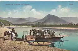 Carte postale montrant une barque à Tchouïozy sur la Katoun avant la construction du pont d'Inya. En arrière-plan se trouve un paysage montagneux verdoyant, ainsi que quelques maisons en bois sur l'autre rive. Cinq personnes et trois chevaux sont déjà sur la barque, tandis que quatre autres sont sur la berge avec leurs deux chevaux. Il y a aussi deux petites pirogues à côté de la barque