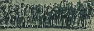 Photo en noir et blanc d'une douzaine d'hommes en tenue de cycliste, debout à côté de leurs vélos qui se tiennent les uns à côté des autres.