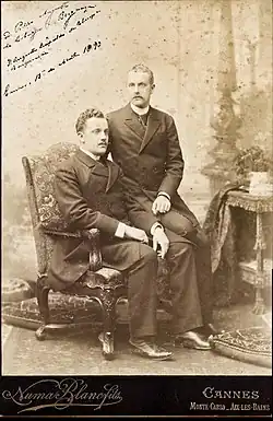 Photo en noir et blanc de deux hommes en habit et redingote, dont l'un est assis sur un fauteuil sur l'accoudoir duquel le second est assis.