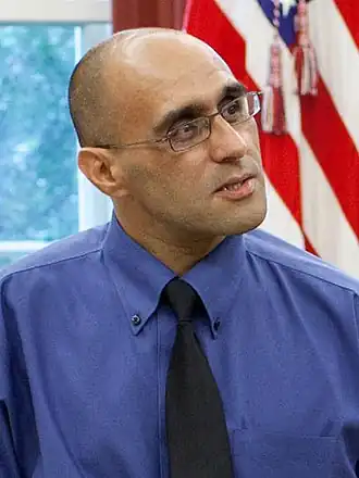 Photographie en couleur, portrait en buste. Un homme chauve avec des lunettes regarde vers la droite, avec le drapeau américain en arrière-plan.