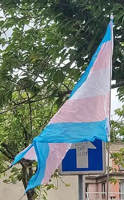 Photographie d'un drapeau de la fierté trans, encore froissé, flottant à côté d'un panneau sens unique tagué sur la flèche "out of the closet"