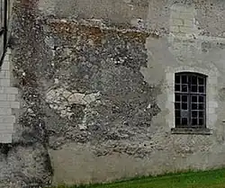 Photographie en couleurs d'un pan de mur au revêtement et parement en partie détruit sur la gauche, une fenêtre à carreau en plein-cintre à droite.