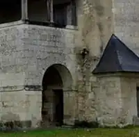 Photographie en couleurs d'une galerie en hauteur surmontant un angle de bâtiment ajourné d'une entrée cintrée, un proche à toiture triangulaire couvert d'un toit fait d'ardoises visible sur la droite.