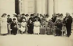 Photographie en noir et blanc. Portrait de groupe d'une vingtaine de femmes accompagnées de quelques hommes : certaines sont assises sur des chaises disposées en rang devant devant une rangée de femmes debout.